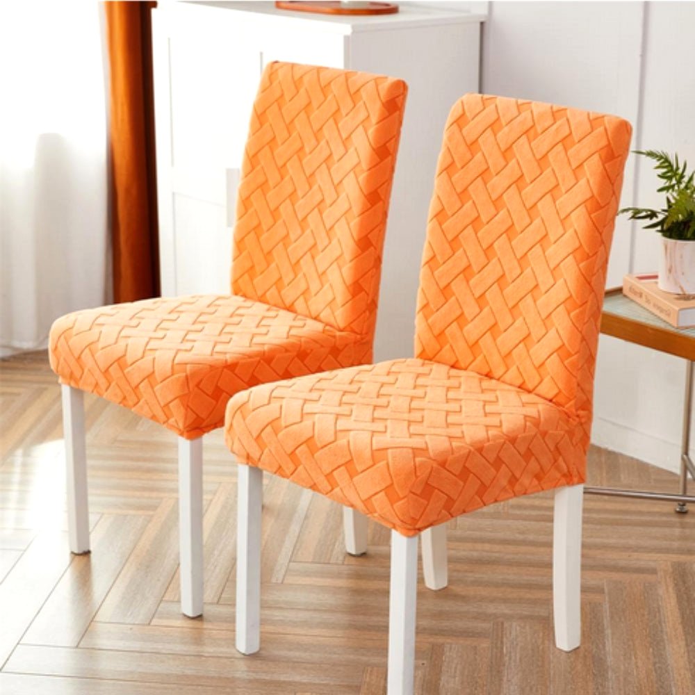 Two dining chairs covered in a vibrant Sunset Orange Haven Weave Jacquard Chair Cover, showcasing the elegant woven pattern. The chairs feature white legs and are placed on a light wood floor in a modern dining setting.