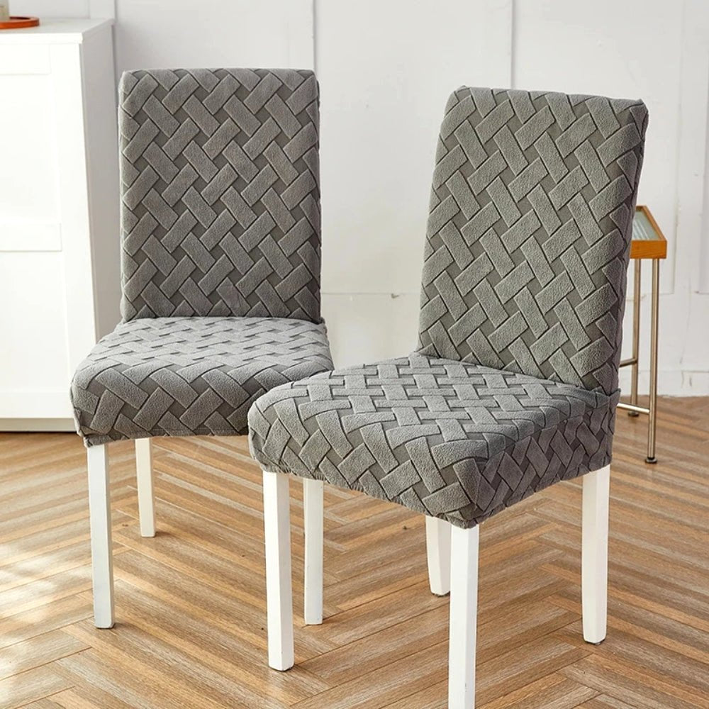 Two dining chairs adorned with a Charcoal Gray Haven Weave Jacquard Chair Cover, highlighting its elegant woven texture. The chairs have white legs and are set in a contemporary room with a light wood floor.